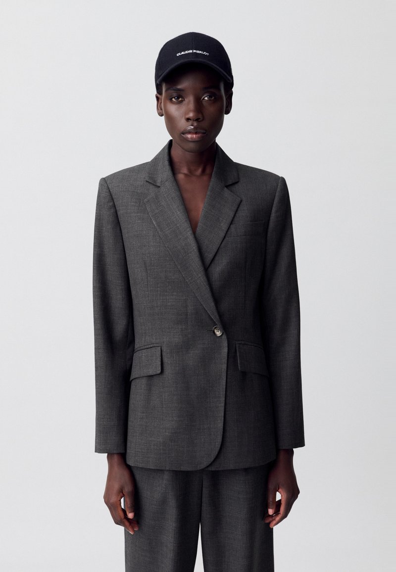 Blazer gris ajustado con dos bolsillos frontales, cierre de un solo botón y solapas afiladas; usado sobre pantalones a juego y una gorra negra.