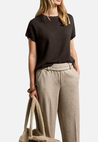 Donna che indossa un maglione corto marrone scuro e pantaloni beige a vita alta, mentre tiene una borsa a tracolla beige con una texture in una mano.
