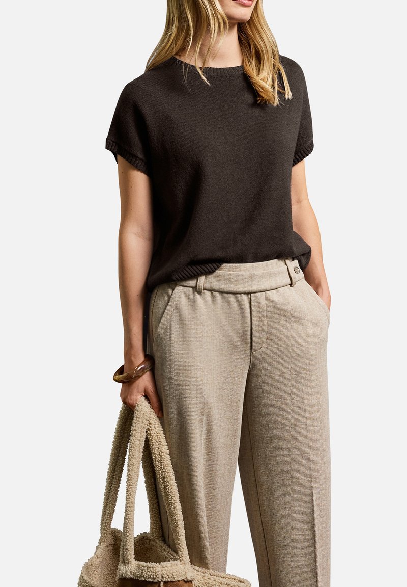 Donna che indossa un maglione corto marrone scuro e pantaloni beige a vita alta, mentre tiene una borsa a tracolla beige con una texture in una mano.