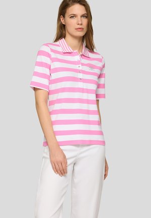 Frau trägt ein pink-weiß horizontal gestreiftes Poloshirt mit kurzen Ärmeln und weiße Hose, steht vor einem einfarbigen Hintergrund.