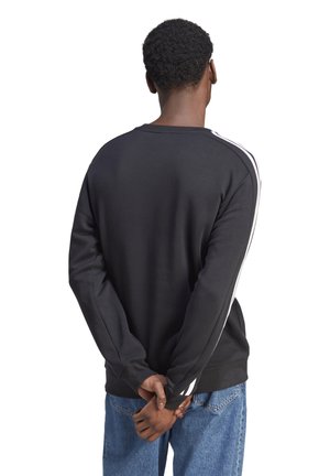 Sudadera negra con franjas laterales blancas, escote redondo, mangas largas y corte relajado, combinada con pantalones de mezclilla azules.