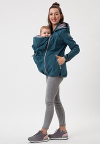 Türkise, mit Reißverschluss und Kapuze versehene Jacke mit einer vorderen Tasche für ein Baby. Das Innere ist grau; sie hat einen lässigen Schnitt und Seitentaschen. Kombiniert mit grauen Jeans und Schuhen.