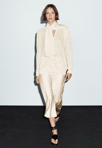 Light beige knit cardigan with scarf, open neck, and buttons, paired with a shiny, beige skirt, and black sandals with ankle straps.