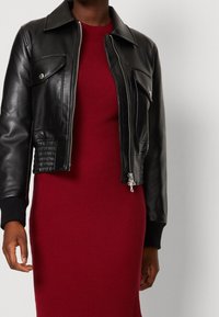 Personne portant une veste en cuir noire avec une fermeture éclair et des boutons argentés sur une robe rouge côtelée, sur un fond uni.