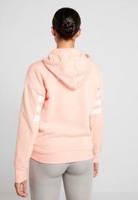 Hoodie rosa chiaro realizzato in tessuto morbido, con cappuccio regolabile e strisce bianche su entrambe le maniche. Texture liscia; design dalla vestibilità rilassata.