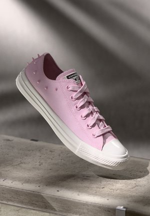 Baskets Converse rose clair avec semelle en caoutchouc blanche et embout blanc, ornées de petites pointes décoratives sur la zone du talon, posées sur une surface en béton.