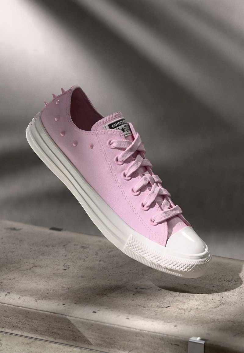 Baskets Converse rose clair avec semelle en caoutchouc blanche et embout blanc, ornées de petites pointes décoratives sur la zone du talon, posées sur une surface en béton.