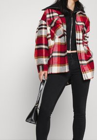 Mujer vestida con una chaqueta de cuadros roja, blanca y marrón sobre un top corto negro y unos vaqueros ajustados negros, sosteniendo un pequeño bolso negro.