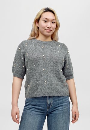 Une femme porte un pull gris à manches courtes orné de perles imitant des perles, avec un jean bleu, sur un fond uni.