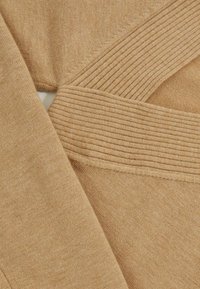 Brązowy dzianinowy materiał o miękkiej fakturze, z akcentami w prążki i ukośnymi detalami szwów. Zbliżenie ukazuje nakładające się warstwy materiału.