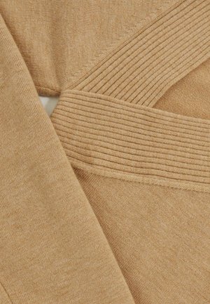 Tan gebreide stof met een zachte textuur, voorzien van geribde accenten en diagonale naaddetails. Close-up view benadrukt de overlappende materiaallaag.