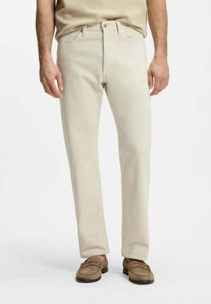 Mann trägt beige gerade geschnittene Hosen und braune Mokassins, steht vor einem schlichten weißen Hintergrund.