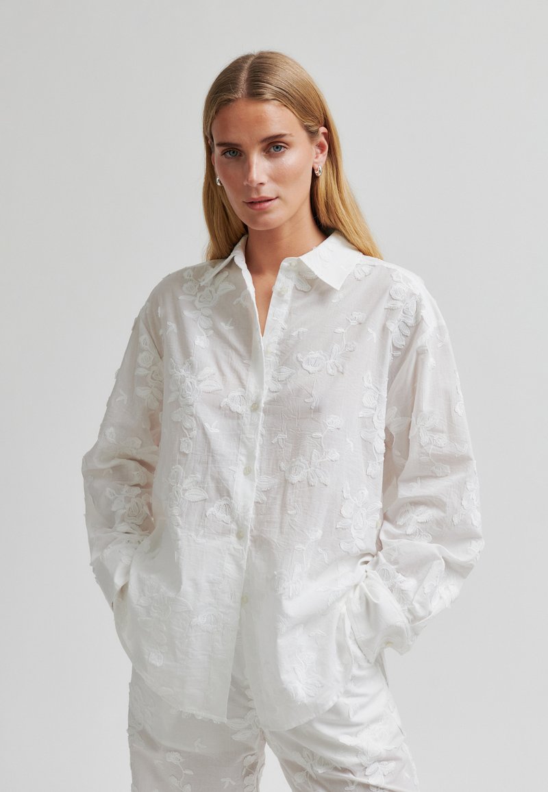 Femme aux longs cheveux blonds portant une chemise boutonnée blanche brodée de fleurs et un pantalon assorti, les mains dans les poches, fond neutre.