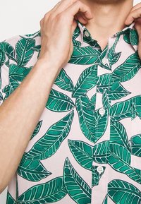 Mani che sistemano il colletto di una camicia rosa con una grande stampa di foglie tropicali verdi e bottoni bianchi sulla tasca anteriore e sulla parte centrale.