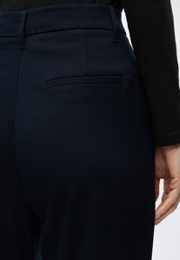Gros plan d'une personne portant un pantalon bleu marine foncé avec une poche passepoilée arrière et un haut noir à manches longues, main reposant sur le côté.