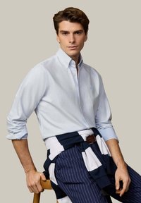 Jeune homme assis sur un tabouret en bois, portant une chemise bleu clair boutonnée, un pantalon rayé bleu marine et un pull bleu marine et blanc noué autour de la taille.