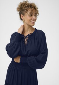 Tumši zilā blūze ar frāzētu kakla izgriezumu, saites detalizāciju un garām pūkainām piedurknēm. Ražota no teksturēta auduma ar elastīgu vidukļa akcentu.