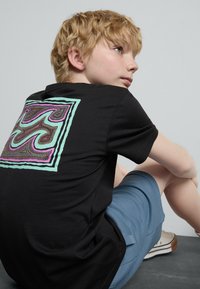 Svart bomulls T-skjorte med et fargerikt grafisk design på baksiden, med svingende linjer i mintgrønn, rosa og gul på en firkantet kant.
