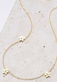 Collana in oro con tre pendenti a forma di fiore bianco, ognuno con uno strass centrale, adagiata su uno sfondo neutro e testurizzato.
