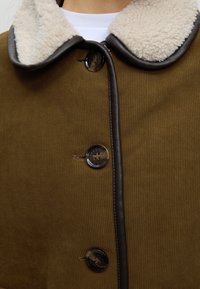 Gros plan d'un manteau marron boutonné avec une bordure sombre et un col en polaire blanche sur une chemise blanche.