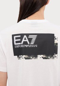 Balta kokvilnas t-kreklu ar melnu grafiku uz muguras, kurā attēlots "EA7 Emporio Armani" un faktūras pikselizēts kamuflāžas raksts.