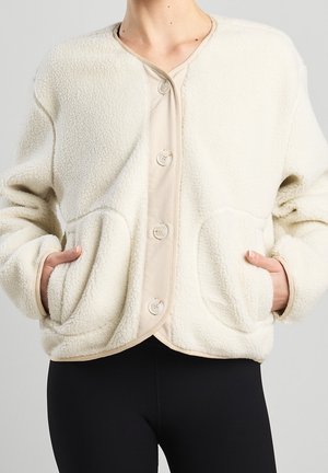 Veste polaire couleur crème à boutons avec grandes poches avant, portée sur une chemise beige, associée à un pantalon noir, mains dans les poches.