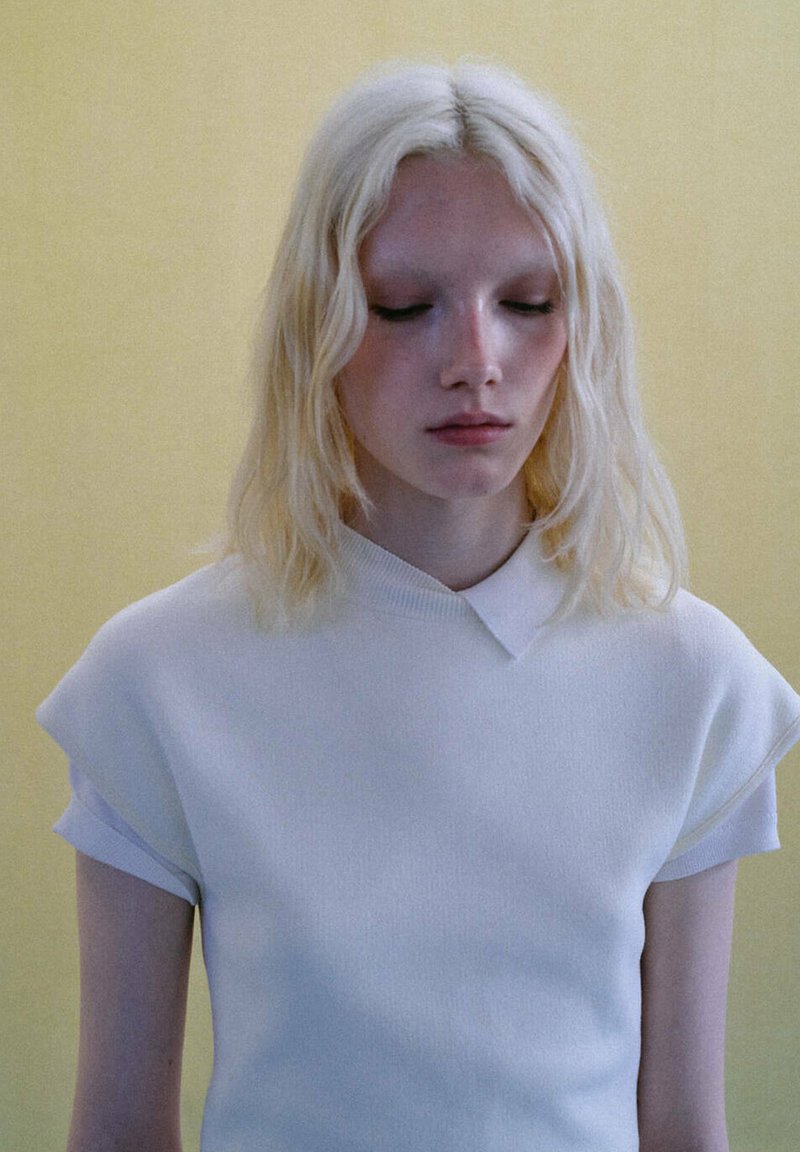 Jeune personne à la peau pâle et aux cheveux blond platine mi-longs, portant un haut blanc à manches courtes sur un fond jaune uni.