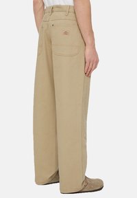 Béžové široké kalhoty vyrobené z odolného materiálu, s velkými zadními kapsami a viditelným šitím. Design má uvolněný, volný střih.