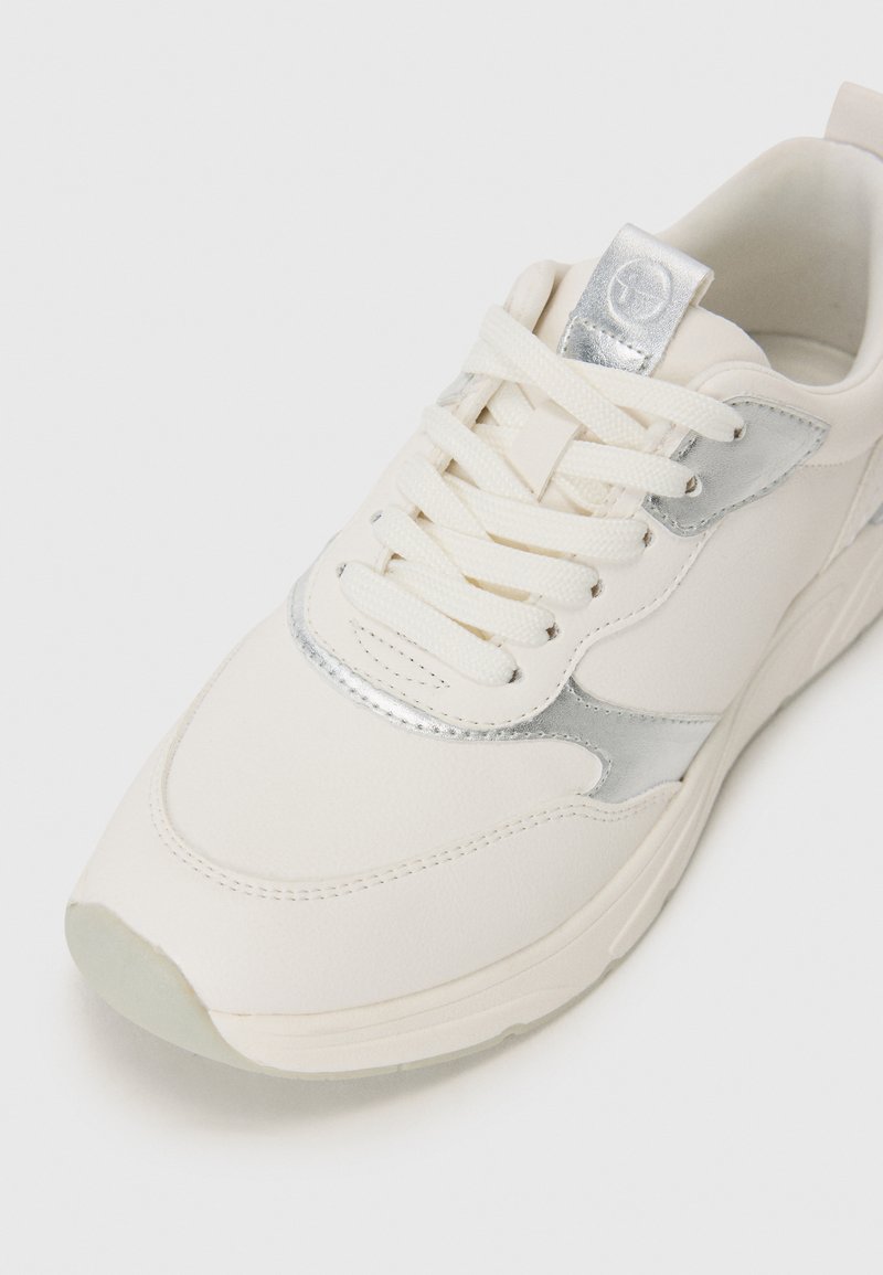 Baskets blanches en cuir lisse, avec des accents métalliques argentés, bout rond et semelle texturée. Présente des lacets plats et une languette rembourrée.