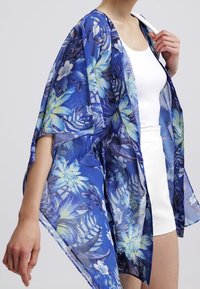Žena v průhledném modrém květinovém kimono přes bílé bezrukávové šaty, drží oděv u límce.
