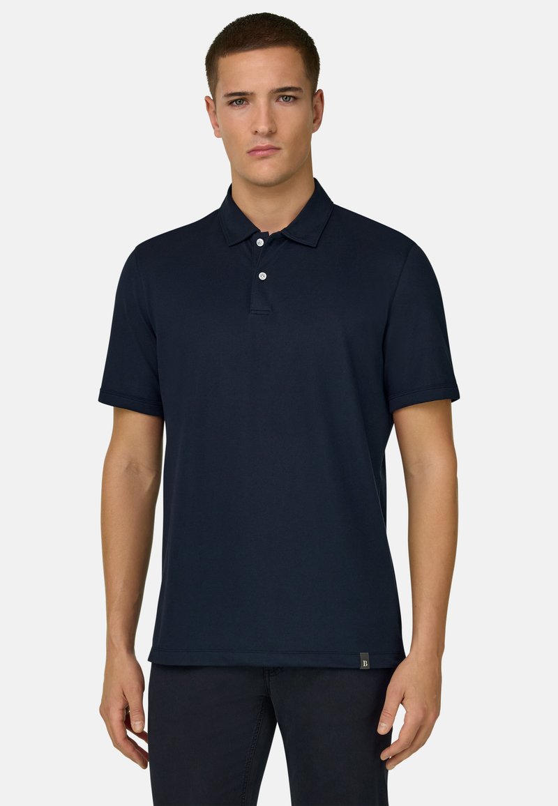 Jeune homme portant un polo uni bleu marine à manches courtes, debout contre un fond blanc, face à l'avant.