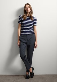 Gestreept donkerblauw en wit fitted top, gecombineerd met donkergrijze op maat gemaakte broek en zwarte instappers, tegen een neutrale muur. Strakke texturen en schone lijnen.