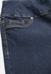 Mörk denimjeans med gula sömmar, en myntficka och en synlig knapp. Tyget har en slät textur med viss blekning.