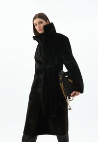 Černý kabát z umělé kožešiny s vysokým límcem, zapínáním na knoflíky a zavázaným pasem, doplněný malou černou taškou s pozlacenými řetízkovými detaily.