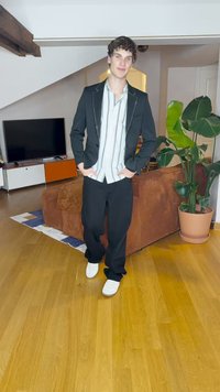 Blazer nero con borchie argento, camicia a righe bianche e blu, pantaloni neri e sneakers bianche. Ambiente interno con piante e mobili.