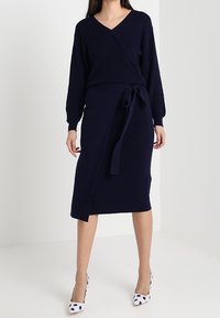 Abito avvolgente in maglia blu navy con maniche lunghe, vita con cintura, orlo fino al ginocchio, abbinato a décolleté bianche con pois neri.