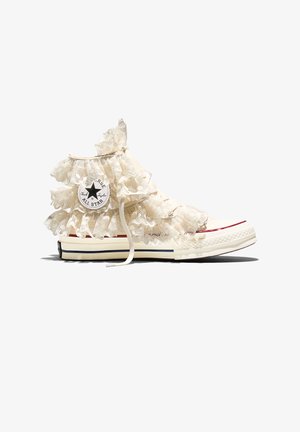 Witte hoge Converse-sneaker met gelaagde roomkleurige kant ruches en zichtbaar merkembleem aan de zijkant, geïsoleerd op een witte achtergrond.