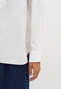 Vit långärmad skjorta med en slät textur, med en avslappnad passform, rundad nertill och knäppta manschetter. Bärs över mörka denimtyg.