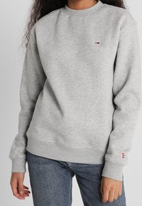 Persona vistiendo un sudadera gris claro con un pequeño logo rojo, blanco y negro en el pecho y la manga, combinada con jeans azules.