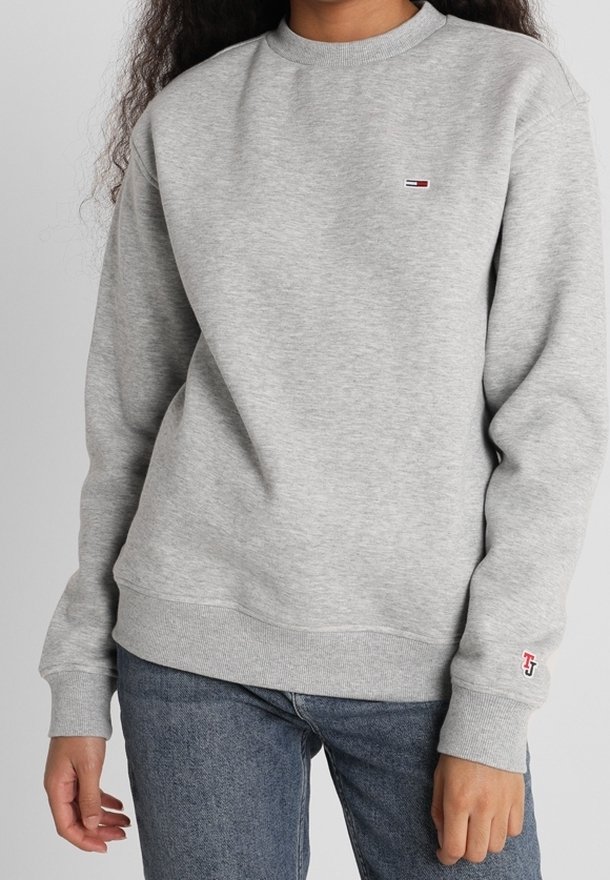 Persona vistiendo un sudadera gris claro con un pequeño logo rojo, blanco y negro en el pecho y la manga, combinada con jeans azules.