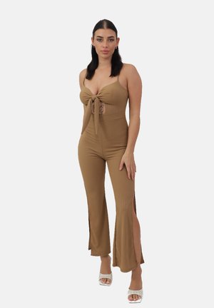 Jumpsuit marrone a coste con un fiocco anteriore, sezione centrale scoperta e gambe svasate con fessure laterali. Indossato con scarpe con tacco trasparenti.
