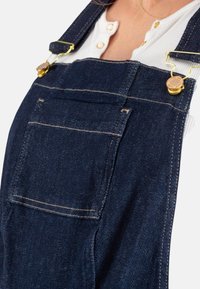 Dunkelblaue Jeanslatzhosen mit goldenen Beschlägen. Verfügt über verstellbare Träger, eine Fronttasche und kontrastierende Nähte für zusätzliche Details.