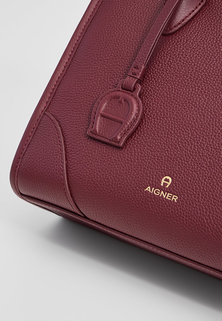 Burgunderfarbene Lederhandtasche mit strukturierter Oberfläche, ausgestattet mit einem goldenen Logo und einem dekorativen Riemen-Detail. Kompakte rechteckige Form.