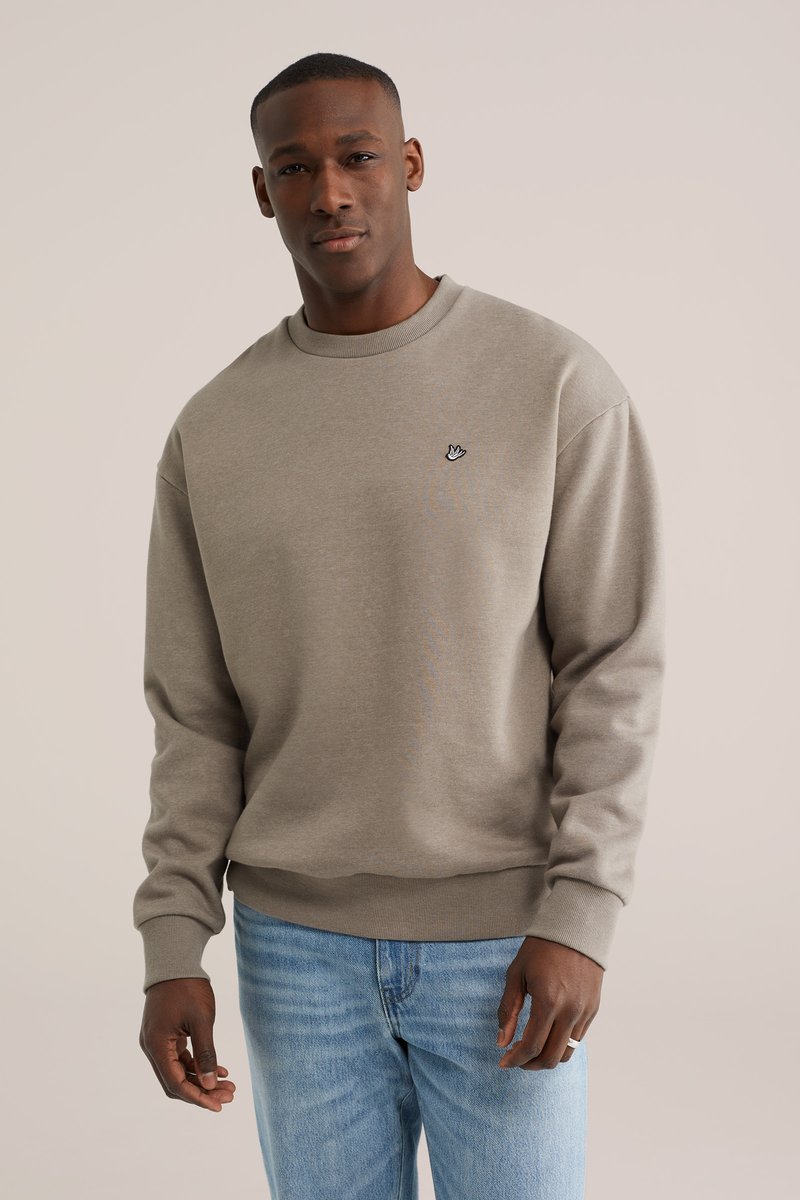 Ung mand iført en beige sweatshirt med rund hals og et lille håndsymbol på brystet samt lyseblå jeans, stående foran en neutral baggrund.