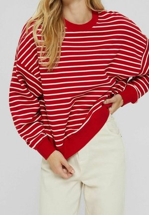 Femme portant un pull à rayures horizontales rouges et blanches avec un pantalon blanc, posant avec une main sur la hanche et l'autre tenant le pull.