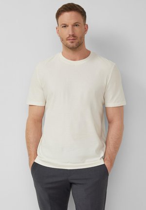 Mann mit kurzem braunem Haar, trägt ein schlichtes off-white kurzärmeliges Hemd und dunkle Hose, steht mit den Händen in den Taschen.