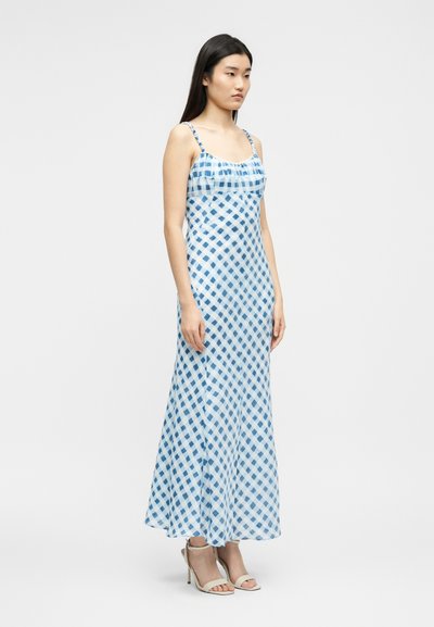Νεαρή γυναίκα που φοράει μάξι φόρεμα χωρίς μανίκια με μπλε και λευκό καρό σχέδιο και μπεζ ψηλοτάκουνα σανδάλια, στέκεται μπροστά σε απλό φόντο.