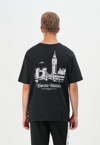 Tricou de bumbac negru cu o ilustrație albă a Podului Tower, a ceasului Big Ben și a unui autobuz cu etaj pe spate. Design casual.