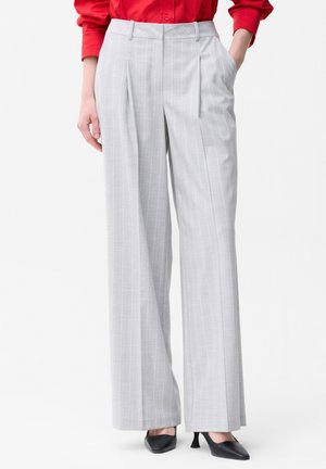 Kobieta ubrana w jasnoszare spodnie z wysokim stanem, szerokimi nogawkami i cienkimi prążkami z plisami z przodu, do tego czarne szpilki z zaostrzonym noskiem i czerwona koszula.