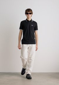 Crna polo majica s bijelim detaljima na ovratniku, uparena s bijelim hlačama i crnim cipelama, nosi je model sa sunčanim naočalama, na jednoličnoj pozadini.